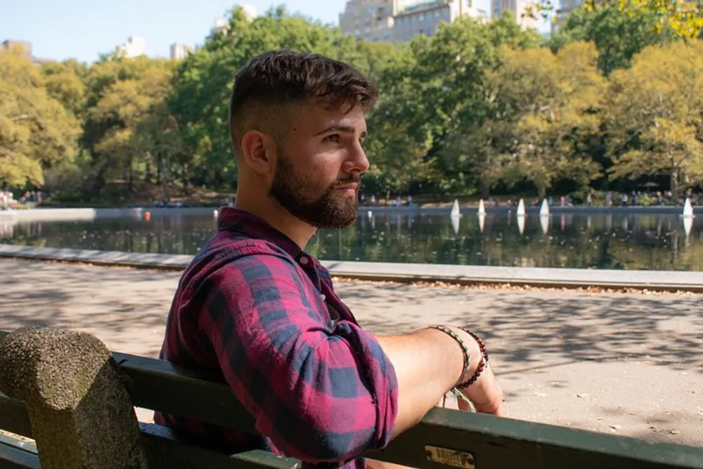 A man with a beard is sitting on a park bench looking to the side with a pond and sailboats in the background