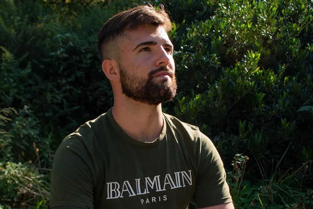 A man with a beard is wearing a green t-shirt and looking to the side with a thoughtful expression with foliage in the background illuminated by sunlight