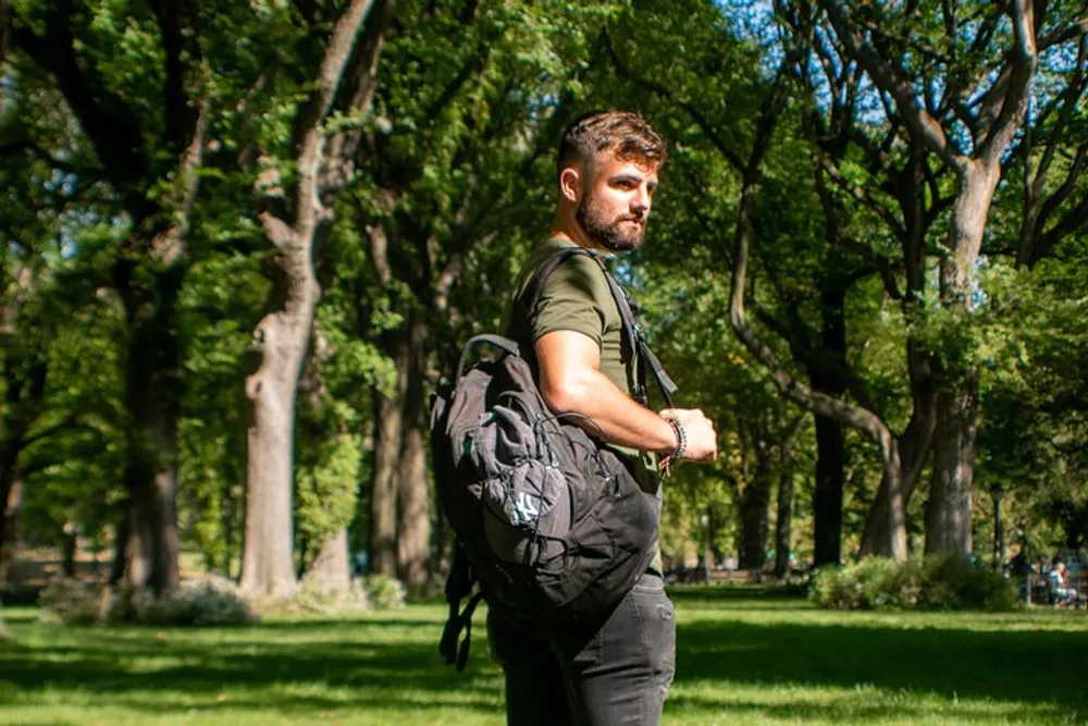 A man with a beard is wearing a green t-shirt and a backpack looking over his shoulder in a lush park setting