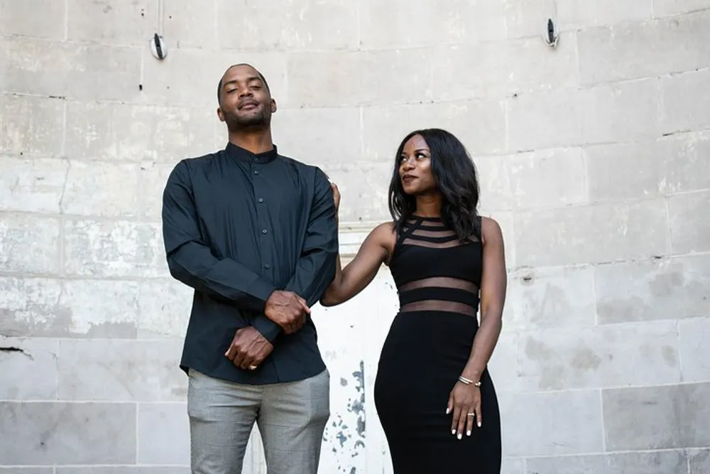 A man and woman are elegantly dressed and holding hands standing against a textured wall with a subtle interaction