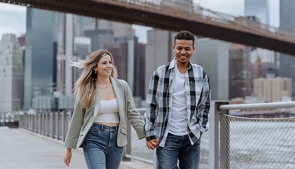 Two people are smiling and holding hands as they walk in an urban setting with a bridge in the background