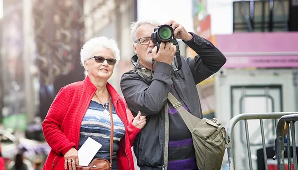 A senior couple appears to be touring a city with the gentleman taking a photograph while the lady stands beside him with a slight smile