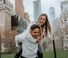 A joyful couple is engaging in a piggyback ride with the woman on the mans back both smiling towards the camera with city buildings and trees in the background