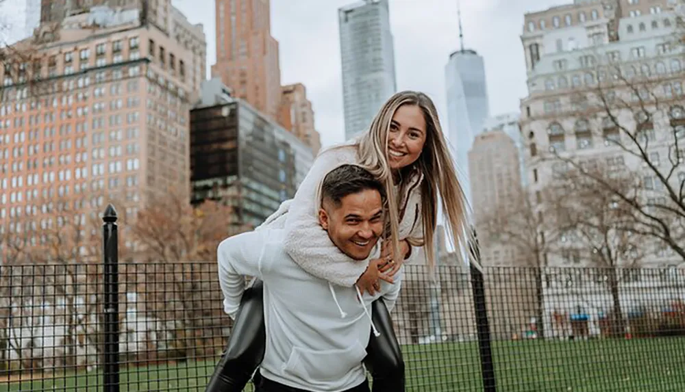 A joyful couple is engaging in a piggyback ride with the woman on the mans back both smiling towards the camera with city buildings and trees in the background