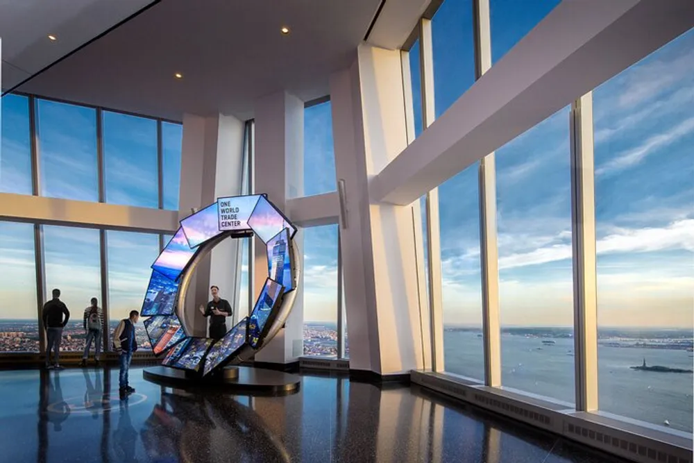 Inside a high-floor observation deck visitors stand near an illuminated pentagonal exhibit with large windows offering a sweeping view of the cityscape and water below