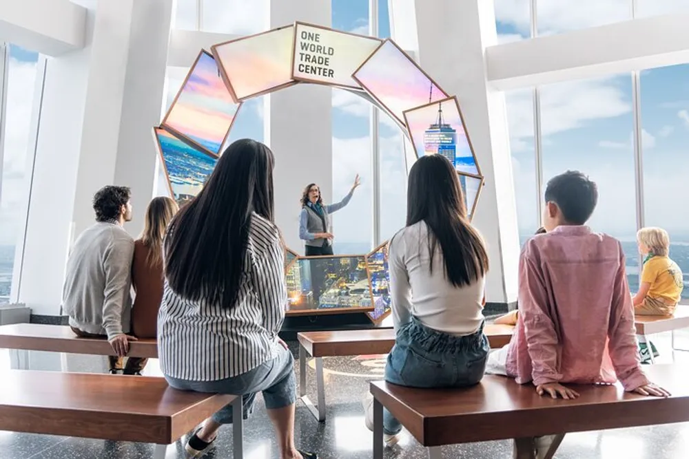 A group of people is attentively listening to a presenter at the One World Trade Center observation deck surrounded by large windows and interactive displays