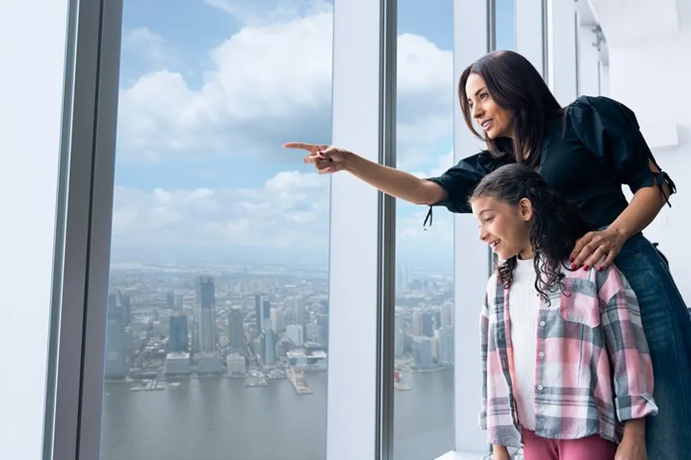 An adult is pointing out something in the distant cityscape through a window to a child both are smiling and seem engaged in the experience