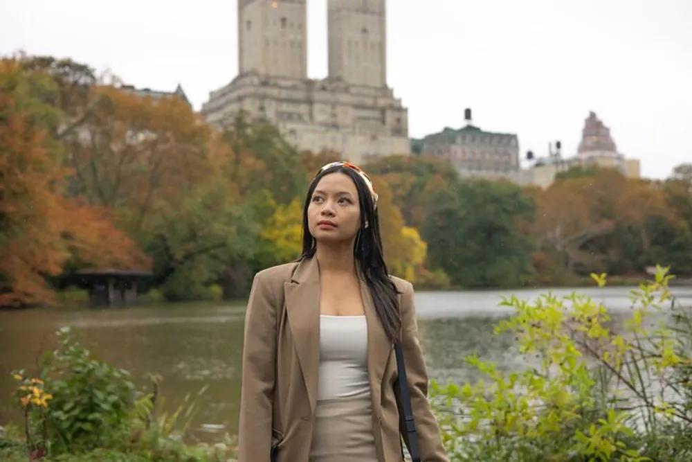 A woman in a beige coat stands contemplatively by a lake in a park with autumn foliage and buildings in the background