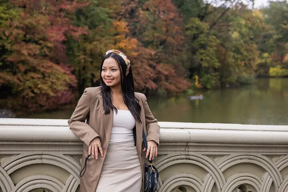 A smiling person is leaning on a balustrade with autumn-colored trees and a pond in the background