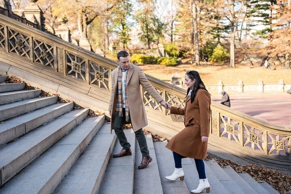 A man and a woman both wearing coats are happily holding hands while descending an outdoor staircase surrounded by autumn foliage