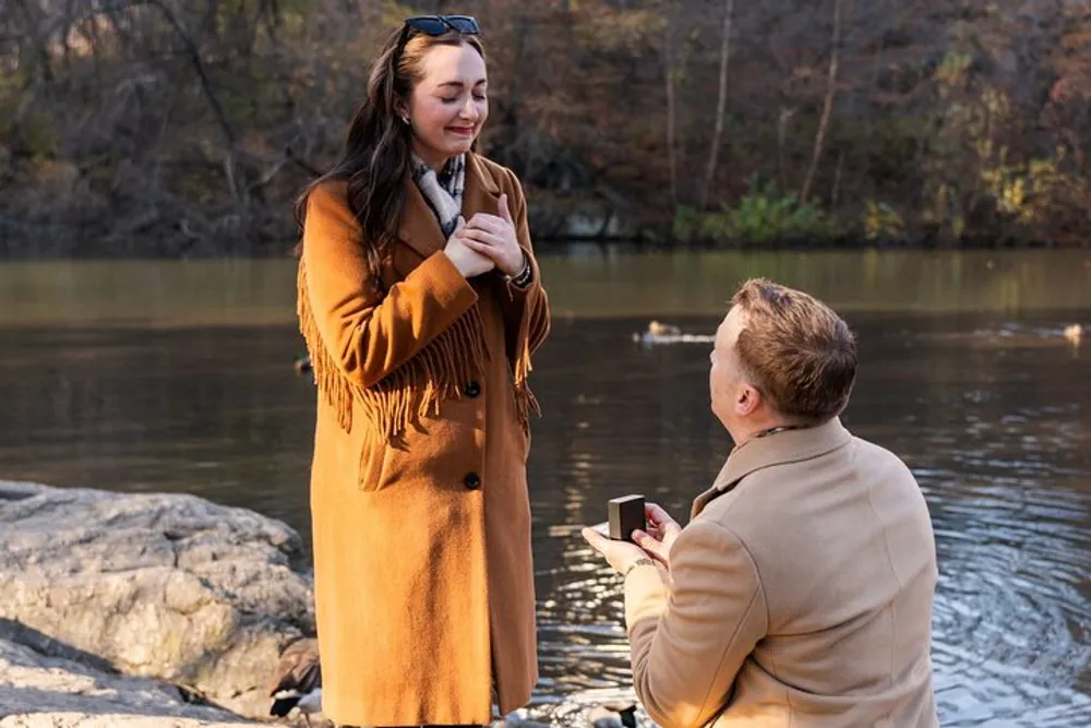 A person is kneeling and holding an open ring box in front of another person who is standing and smiling in what appears to be a marriage proposal near a body of water with ducks in the background