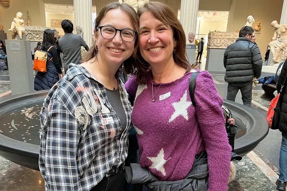 Two smiling individuals are posing together for a photo in a classical museum setting with sculptures and visitors in the background