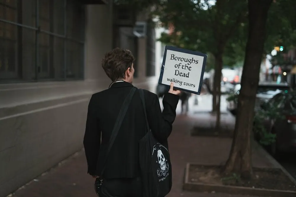 A person in a dark suit holding a sign that reads Boroughs of the Dead Walking Tours is walking down a street