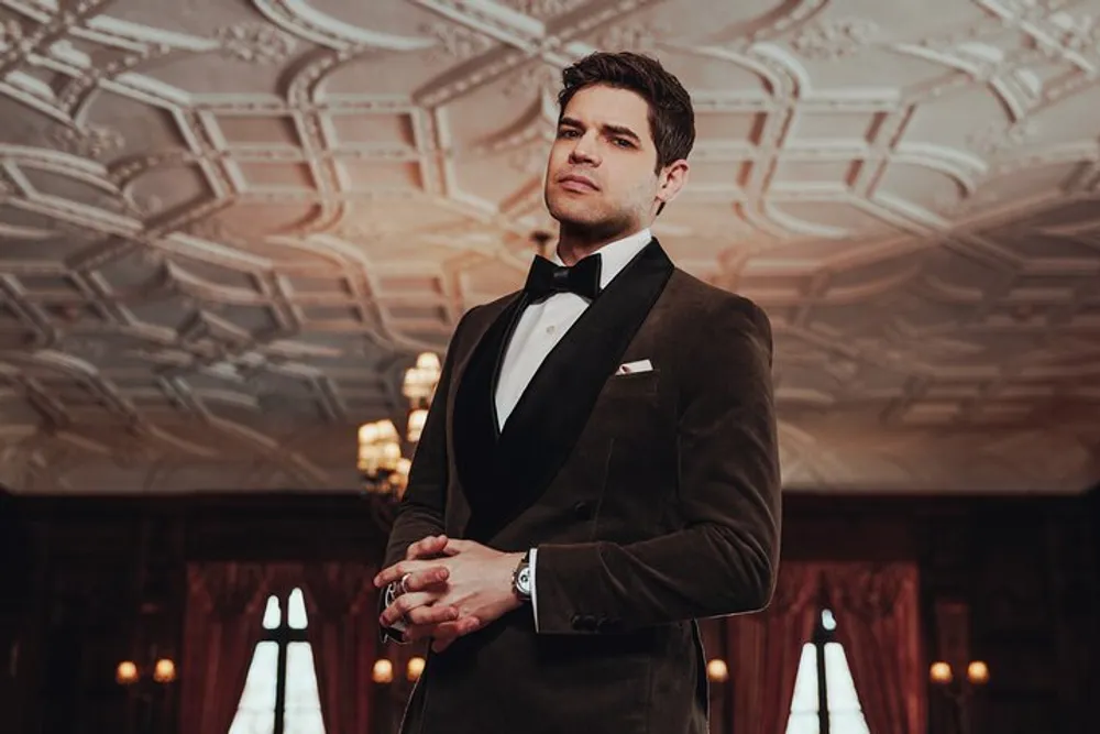 A confident individual in a tuxedo stands in an elegant room with an ornate ceiling