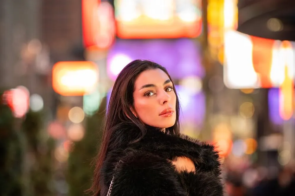 A woman in a black furry jacket poses for a portrait against the vibrant backdrop of illuminated city billboards at night