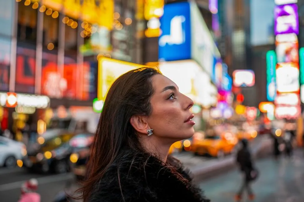 A woman is looking upwards with a contemplative expression against a vibrant backdrop of glowing city billboards and signs at dusk