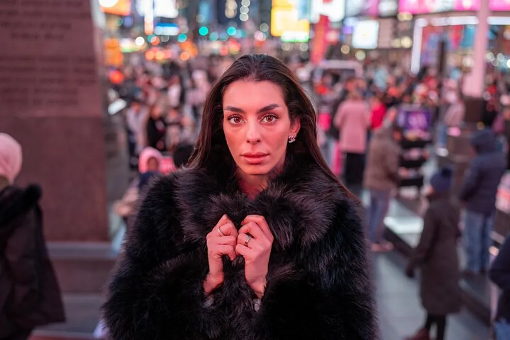 A woman wearing a black fur coat stands in focus against the vibrant backdrop of a busy brightly lit urban night scene