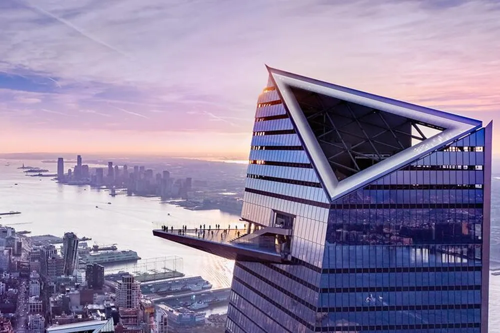 The image shows a striking skyscraper with a distinctive angled top and an exterior observation deck overlooking a cityscape and body of water at sunset