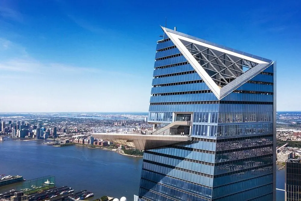 The image shows a distinctive modern skyscraper with a triangular framing at the top overlooking a river and a cityscape under a clear blue sky