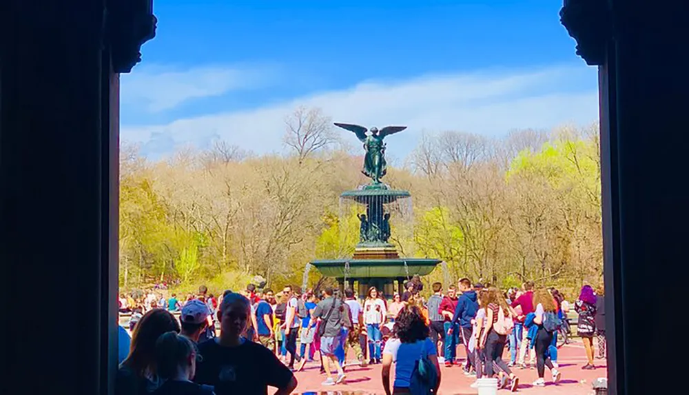 The image offers a view from a shaded area onto a sunny scene where people gather around a fountain topped with an angelic figure possibly in a park setting