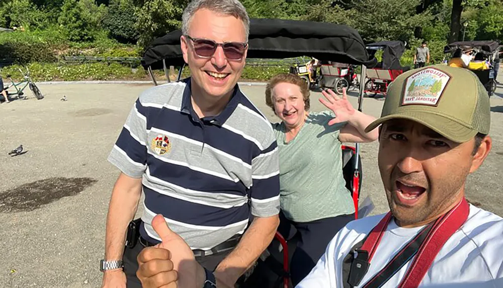 Three people are smiling for a selfie with the person in the foreground giving a thumbs up and the two in the background seated on a park bench waving at the camera