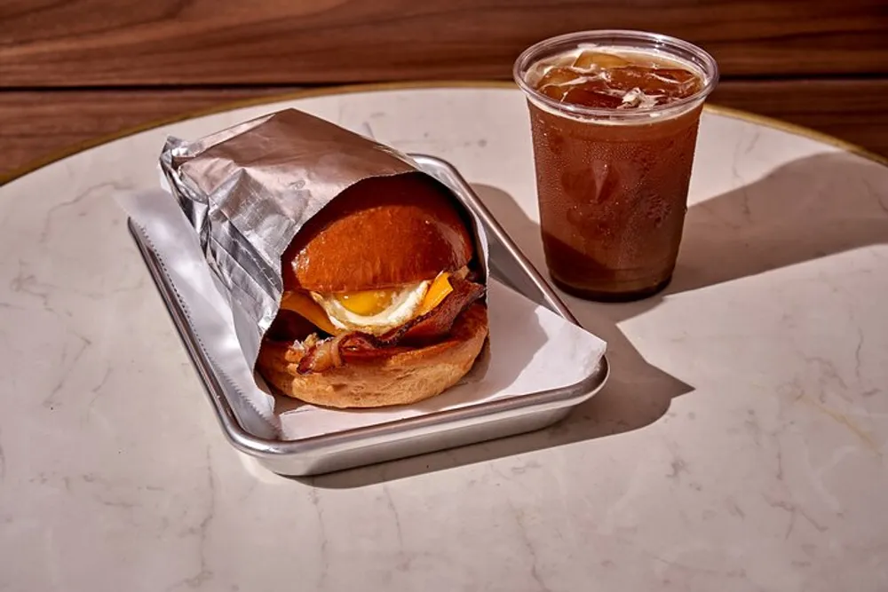 A breakfast sandwich with egg and bacon is partially wrapped in foil on a tray next to a cup of iced coffee