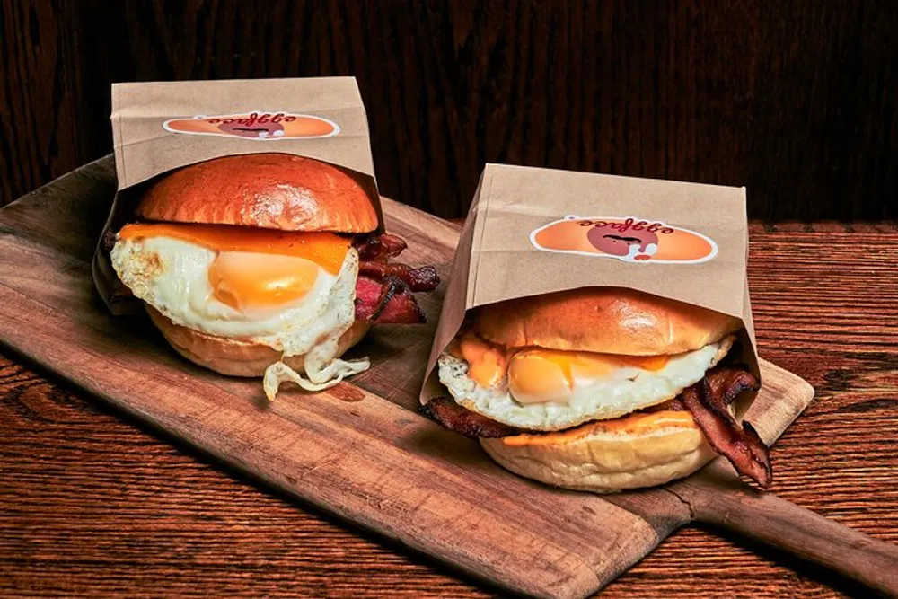 Two breakfast sandwiches with bacon and fried eggs are displayed on a wooden board with branding on paper sleeves