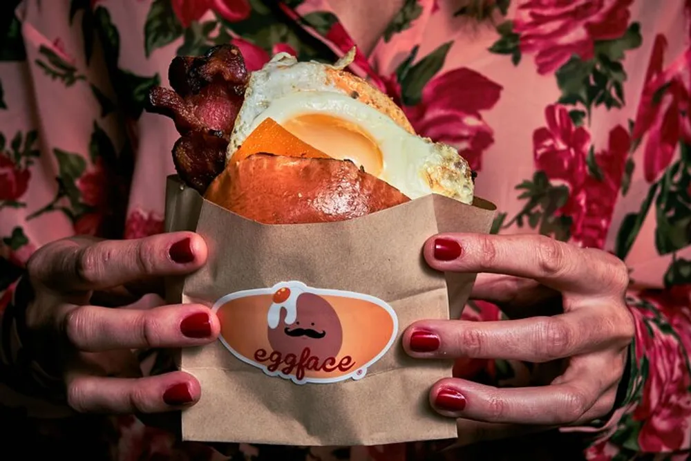 A person is holding a breakfast sandwich with bacon and a fried egg wrapped in a paper holder with the logo eggface