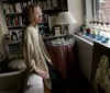 A woman stands contemplatively in a cozy book-filled room with an elaborate collection of glassware displayed on a windowsill