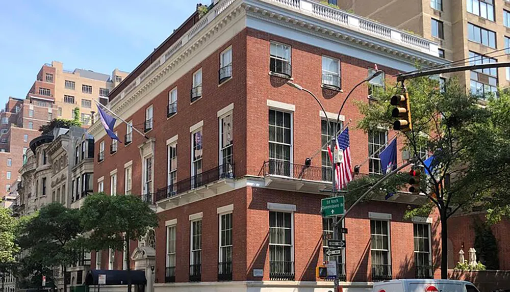 The image shows a stately brick building adorned with multiple flags situated on a street corner with traffic lights and greenery in an urban setting