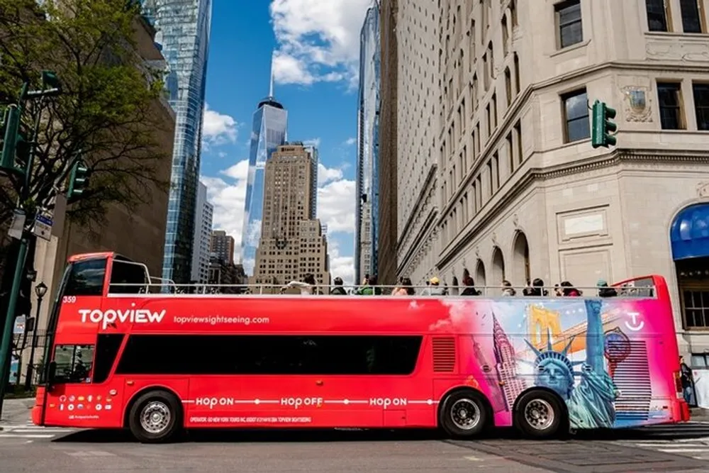 A vibrant red double-decker sightseeing bus is parked on a city street with skyscrapers and partly cloudy skies in the background