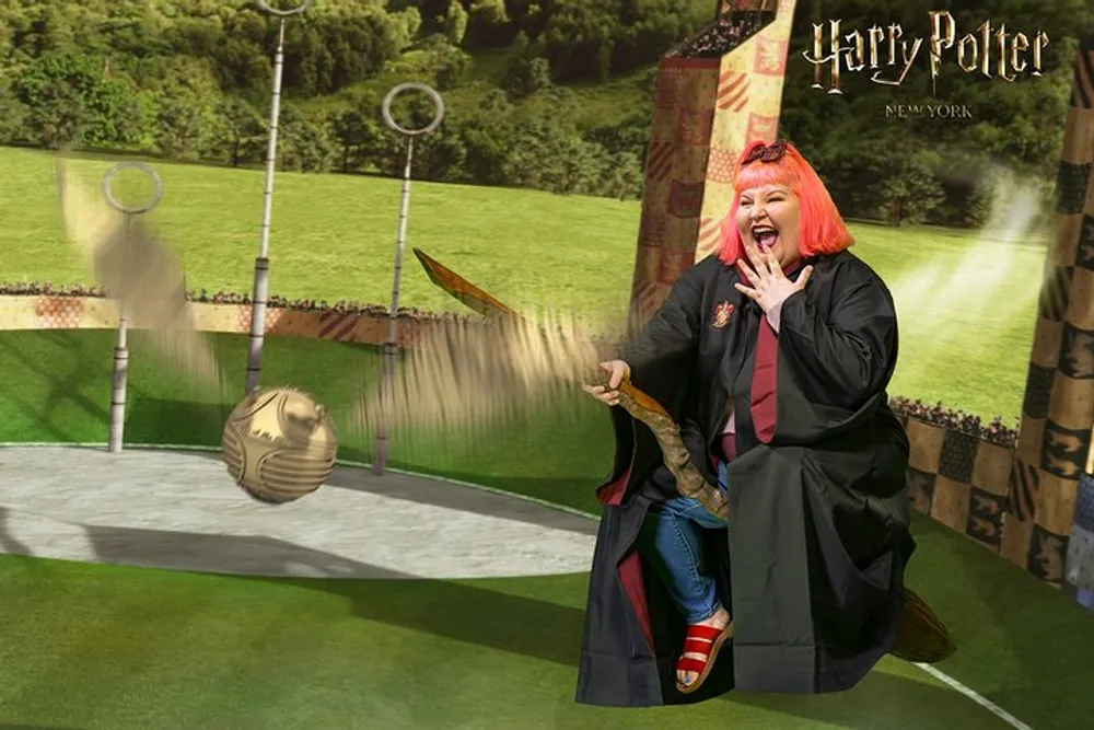 A person is posing joyfully with a broomstick on a Harry Potter-themed Quidditch pitch backdrop as if they are playing the magical sport