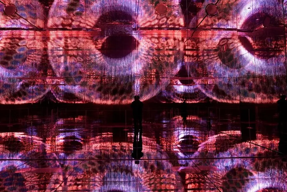 A person stands silhouetted against an immersive vibrant digital art installation featuring a pattern of large eyes on display surfaces