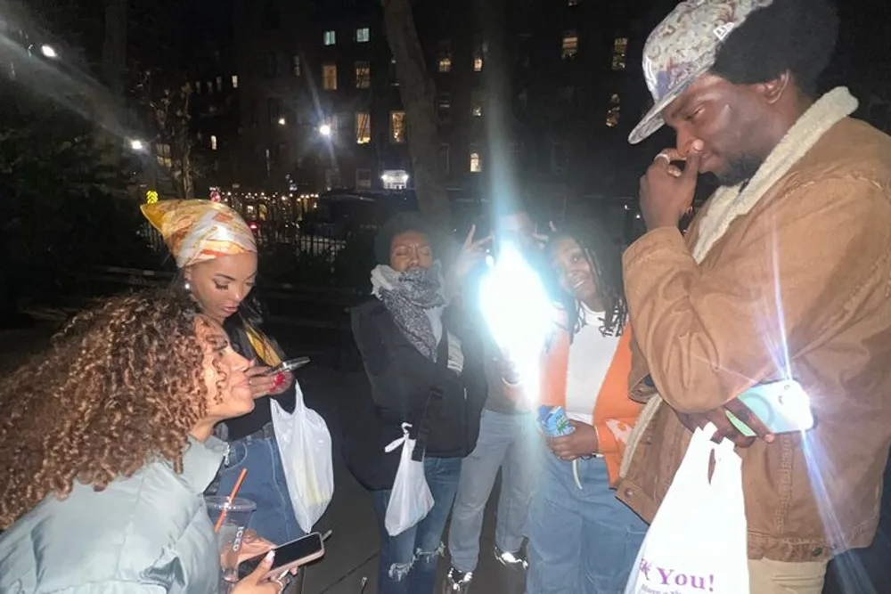 A group of people are gathered at night with one person shining a bright light while others look at their phones or into the distance creating a casual social scene