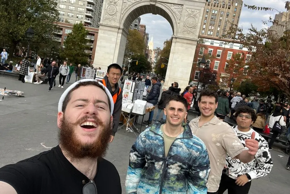 Four friends are taking a smiling selfie with the Washington Square Arch in the background on a lively day