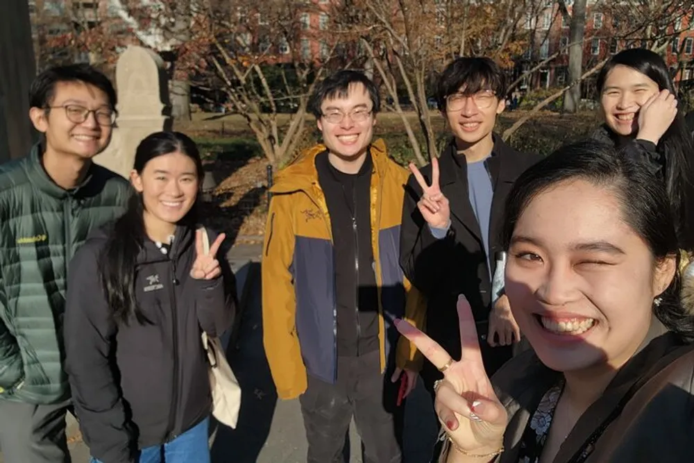 A group of happy young adults poses for a selfie in a sunny outdoor setting with several of them making peace sign gestures