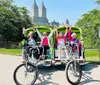 A group of people is cheerfully posing on a pedal-powered rickshaw-like vehicle in a park with tall buildings in the background