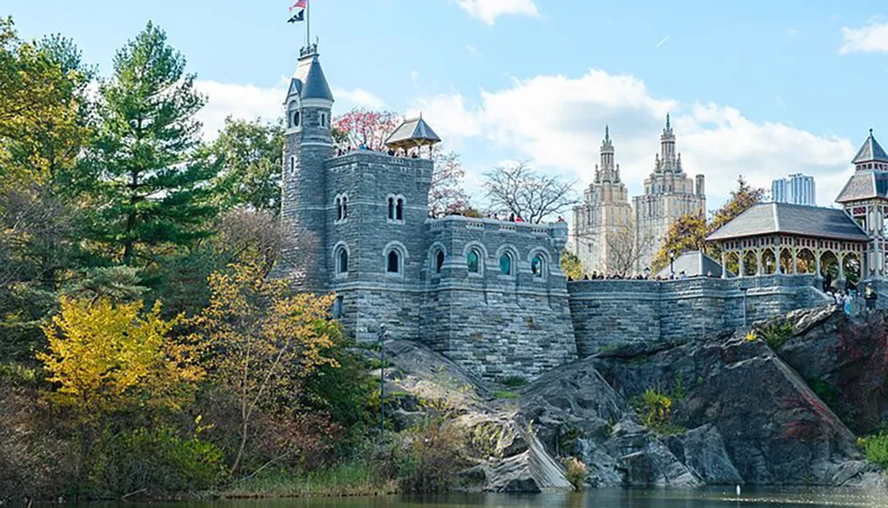 The image features a stone castle-like structure with turrets overlooking a body of water set against a backdrop of autumn foliage and city high-rises