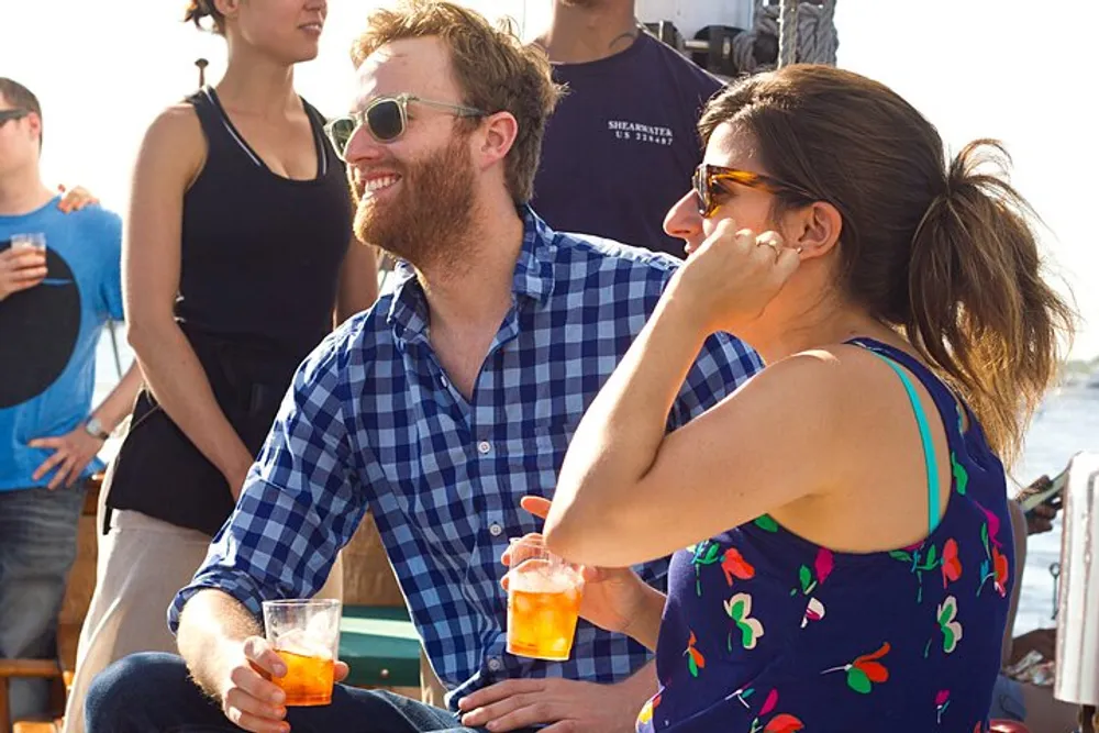 A smiling man in a blue plaid shirt and sunglasses sits next to a woman in a colorful tank top both holding orange drinks in a lively gathering with other people around them