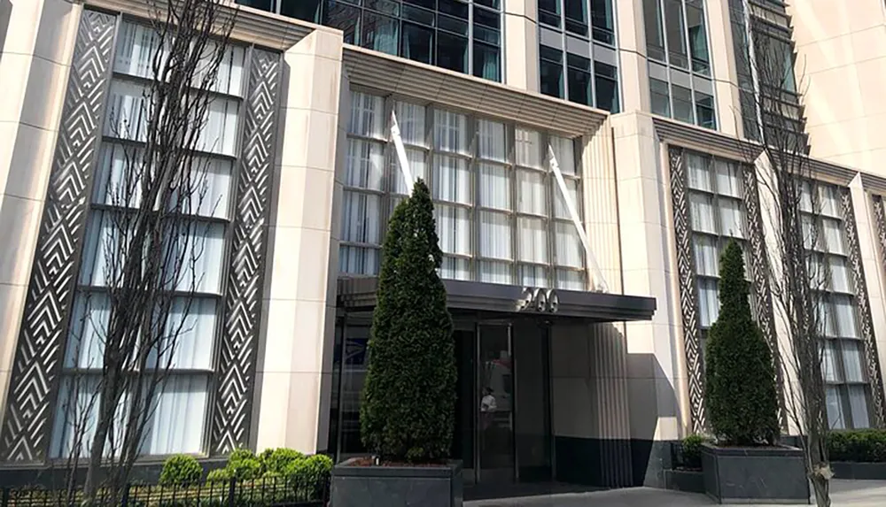 The image shows the exterior of a modern building with distinctive zigzag patterns on its facade large windows and manicured conical topiary trees framing a central entrance