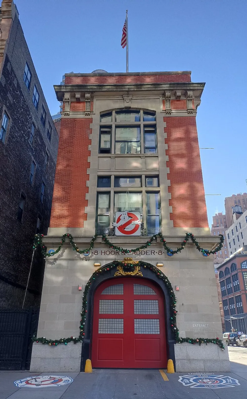 The image shows the exterior of a firehouse adorned with Ghostbusters logo and holiday decorations under a clear blue sky