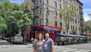 A man and a woman are smiling for a photo on a tree-lined street with a classic urban building in the background, featuring a ground-floor restaurant and an outdoor dining area.