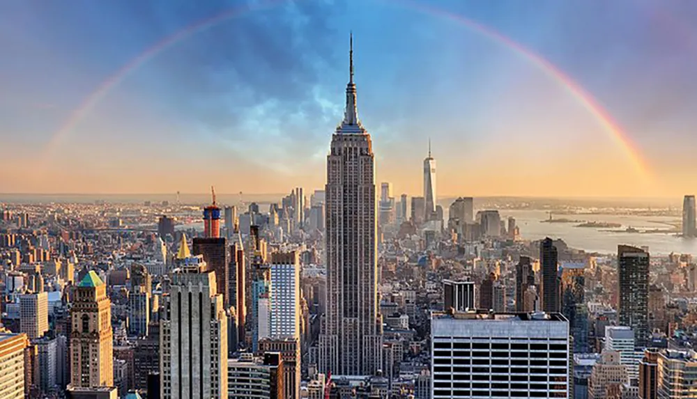 A panoramic view of New York Citys skyline with a prominent view of the Empire State Building under a wide rainbow arching across the scene
