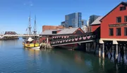 The image features the Boston Tea Party Ships & Museum with a replica of a historical ship moored alongside it, set against the backdrop of modern buildings under a clear blue sky.
