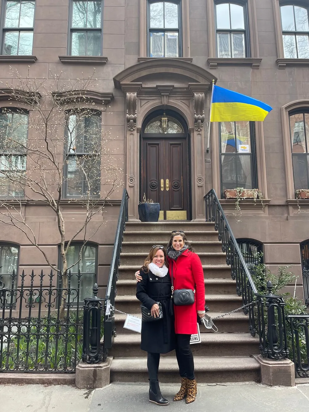 Two smiling women are standing on the steps of a brownstone building under a Ukrainian flag