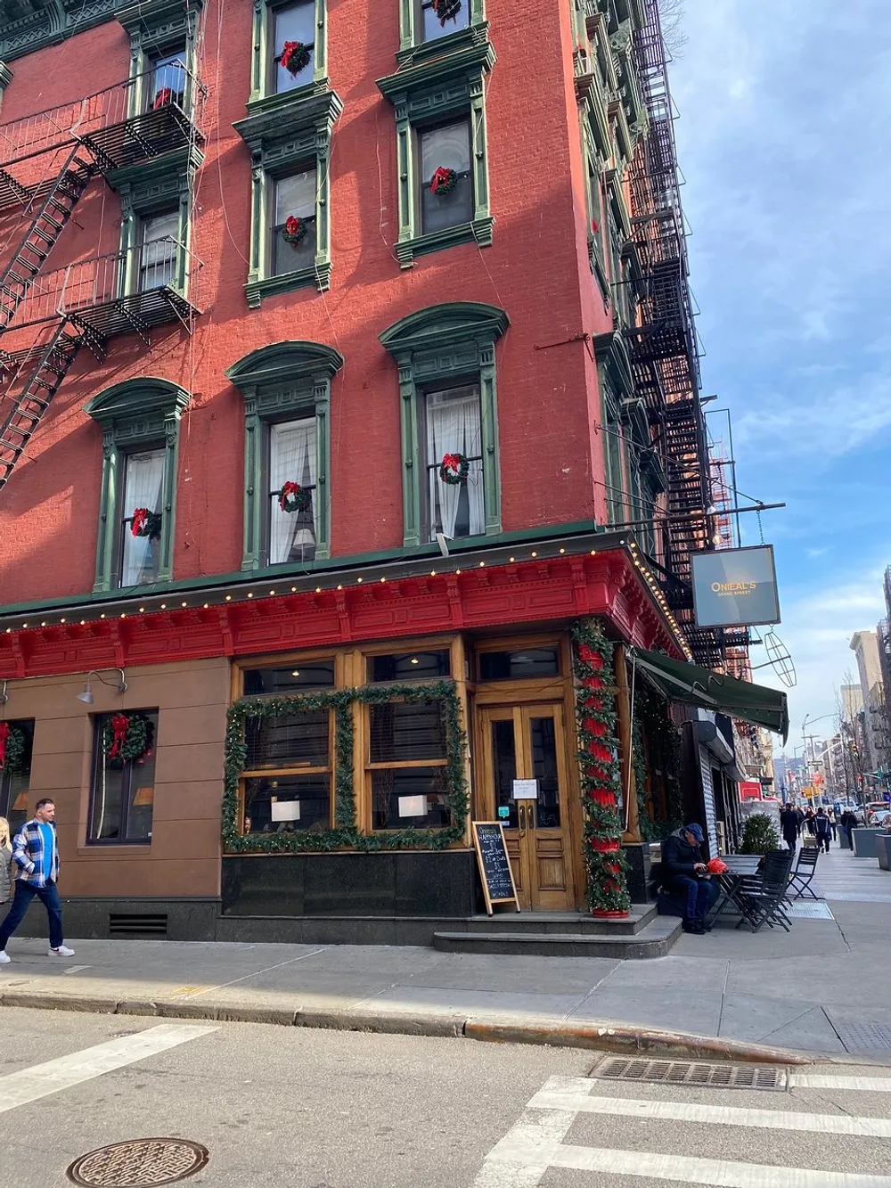 The image shows a festively decorated red brick corner building with green window frames and a fire escape likely a restaurant or bar with people walking by on an urban sunny day