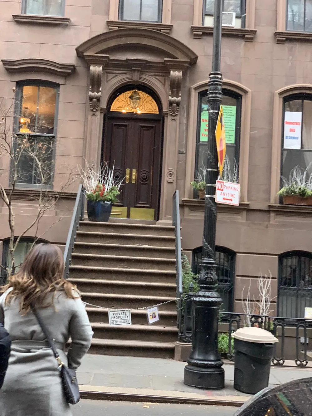 The image shows a woman walking past a brownstone building with a set of stairs leading to an ornamental arched doorway with signs around indicating No Parking Anytime and Private Property No Trespassing