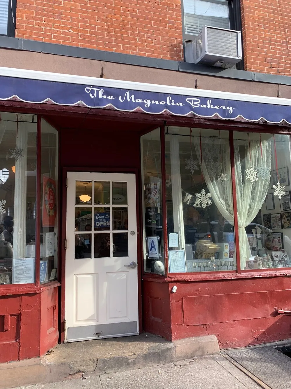 The image shows the exterior of The Magnolia Bakery with a classic red and navy storefront complete with a white door lace-curtained windows and an Open sign suggesting its ready for business