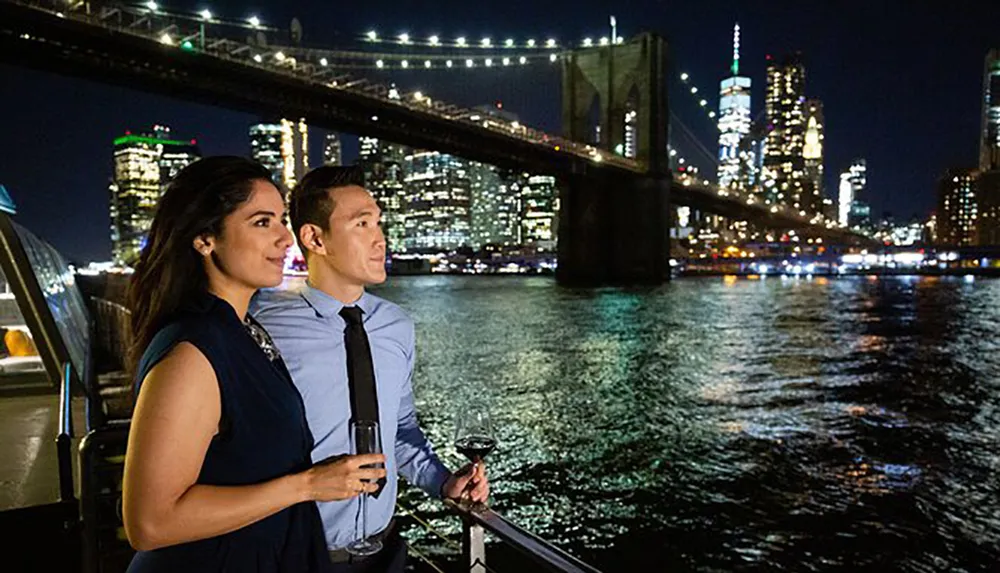 A couple enjoys a nighttime view of a lit-up cityscape with a prominent bridge in the background while holding glasses of wine