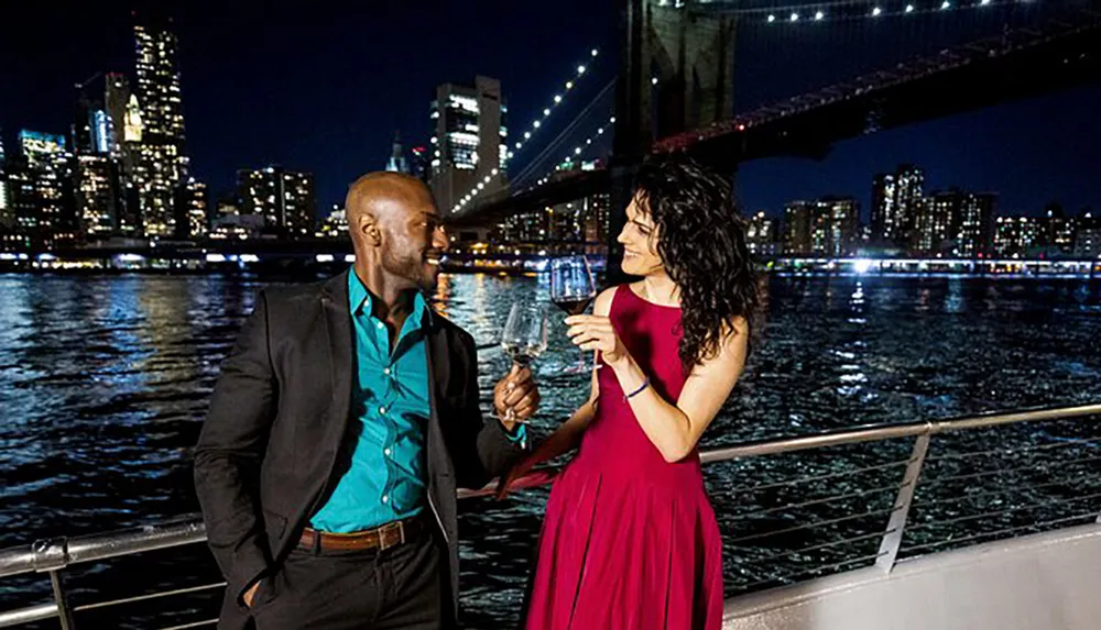 A man and a woman dressed elegantly are sharing a toast with wine glasses against a backdrop of a nighttime city skyline and lit-up bridge
